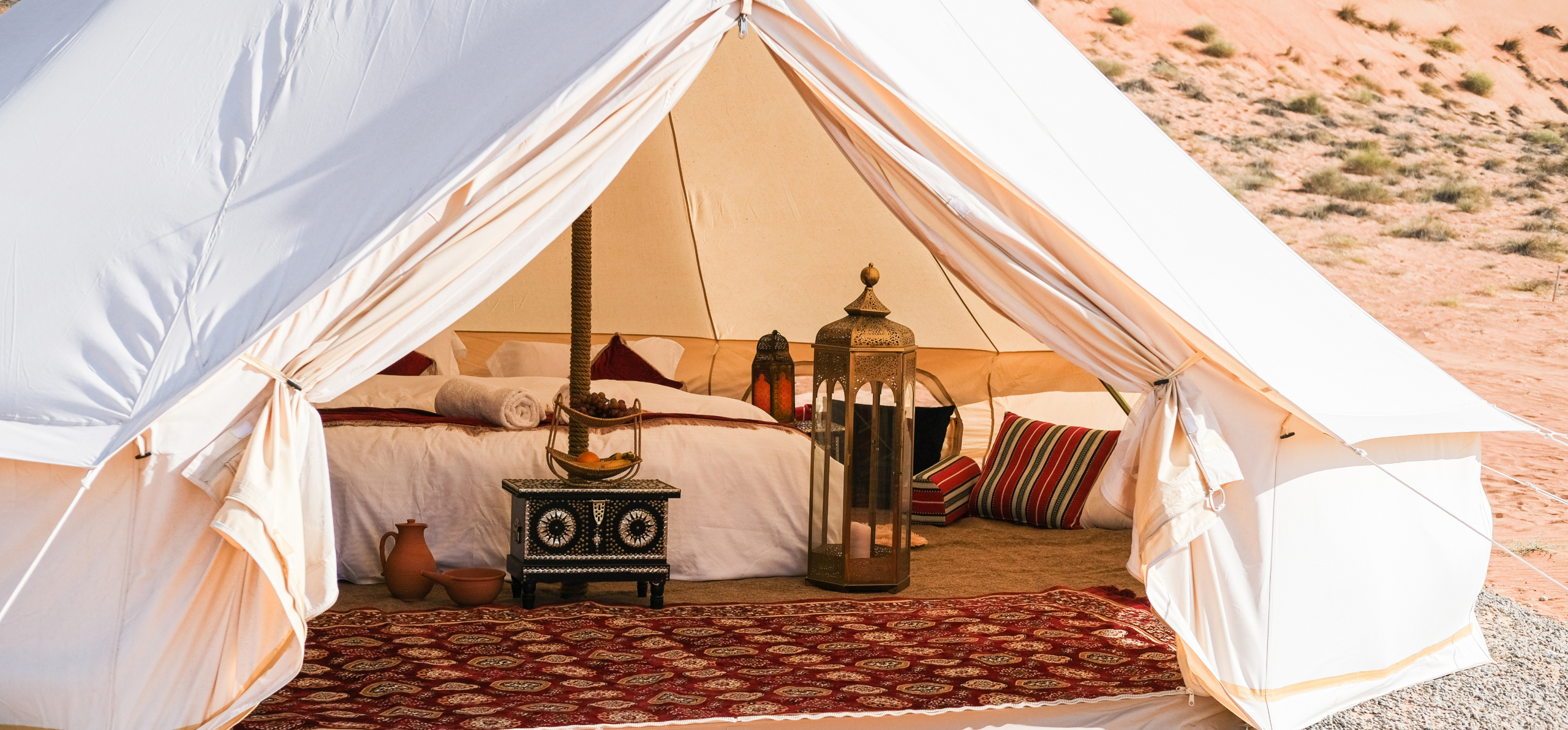 Close photo of spacious white tent in a desert setting. Double bed, red carpet and rich decorative items are visible inside the tent.